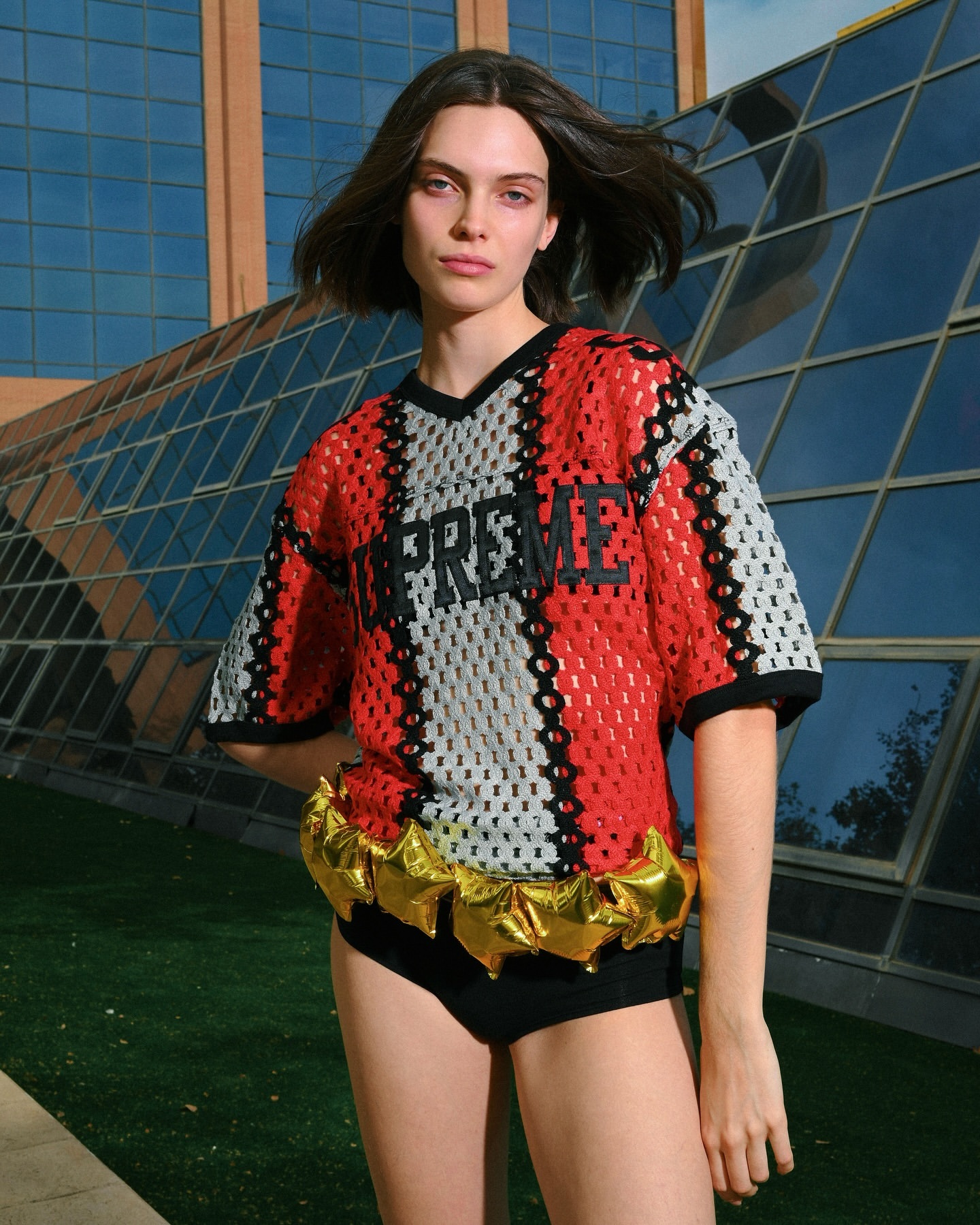 A young woman with dark hair poses outdoors in a red and gray mesh top, and black bottoms near a glass-fronted building.