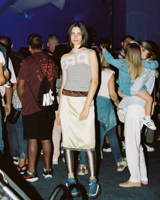 A young adult fashion model poses in a crowded indoor setting wearing a gray tank top and cream colored skirt with metallic tights.