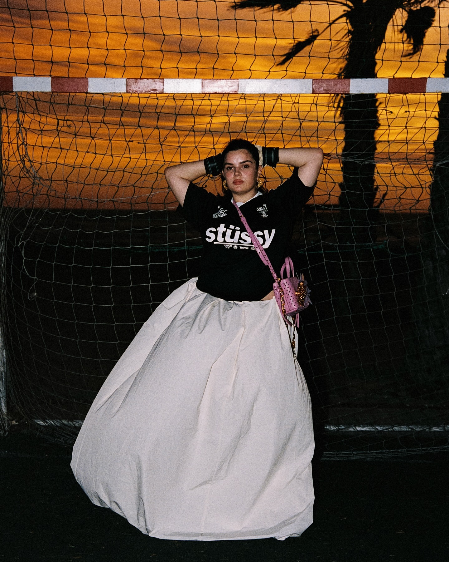 Woman in a Stüssy jersey and white skirt posing in front of a soccer goal at sunset with palm tree shadows, fashion editorial.