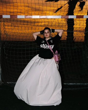 Woman in a Stüssy jersey and white skirt posing in front of a soccer goal at sunset with palm tree shadows, fashion editorial.