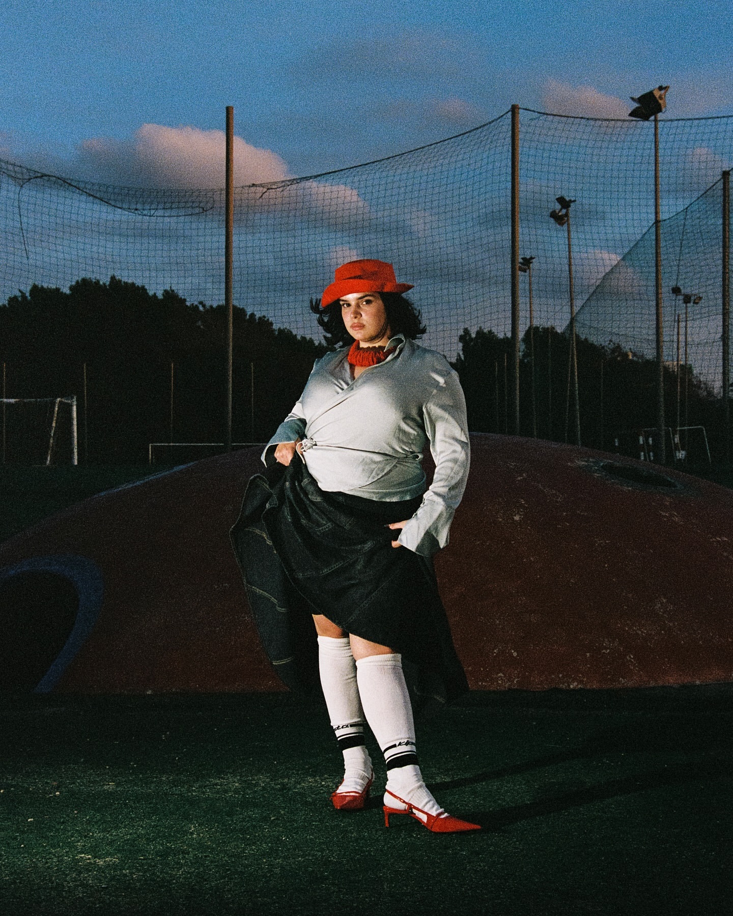 Woman in red hat and heels poses in a sporty outfit on a green field with a soccer net in the background under a cloudy sky.