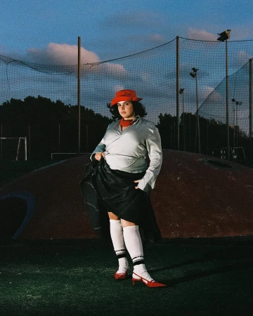 Woman in red hat and heels poses in a sporty outfit on a green field with a soccer net in the background under a cloudy sky.