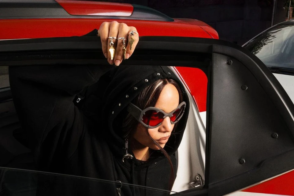 Woman in hooded jacket and sunglasses leans out of a car window with silver rings and key charms on her fingers in an urban scene.