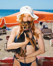 Red-haired woman wearing a white bucket hat and blue bikini holding a small dog on a sunny sandy beach by the ocean.