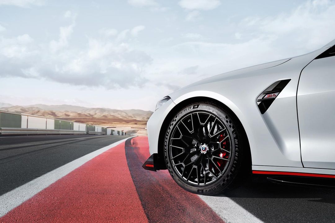 White sports car parked on a racetrack with black wheels, red brake calipers, and a mountain range in the background on a cloudy day.