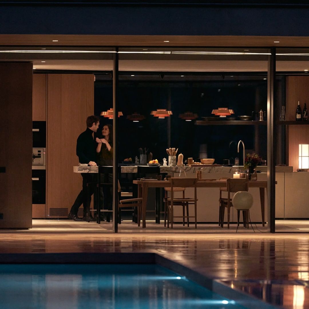 Couple in modern kitchen with pool view at night. Man and woman stand near counter of luxury home with contemporary pendant lighting.