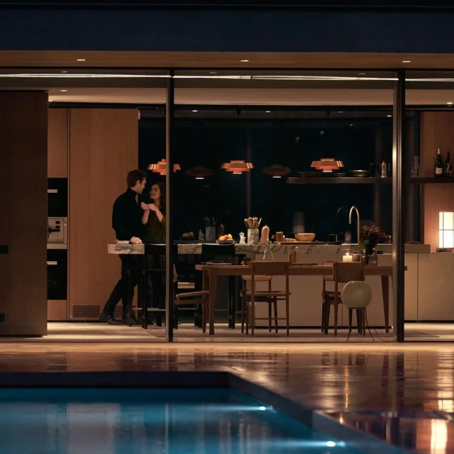 Couple in modern kitchen with pool view at night. Man and woman stand near counter of luxury home with contemporary pendant lighting.