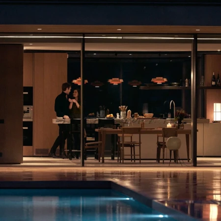Couple in modern kitchen with pool view at night. Man and woman stand near counter of luxury home with contemporary pendant lighting.