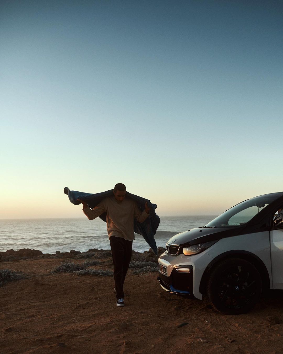 Man walks along the beach at sunset carrying a blanket next to a white BMW i3 electric car with ocean and sky in the background.