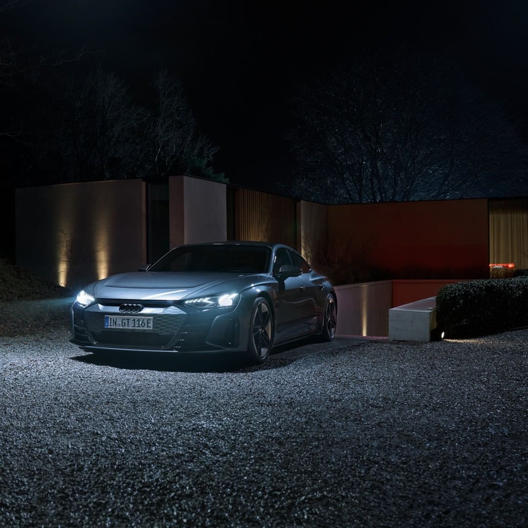 Sleek gray Audi e-tron GT electric car parked on a gravel driveway in front of a modern house at night with its headlights illuminated.