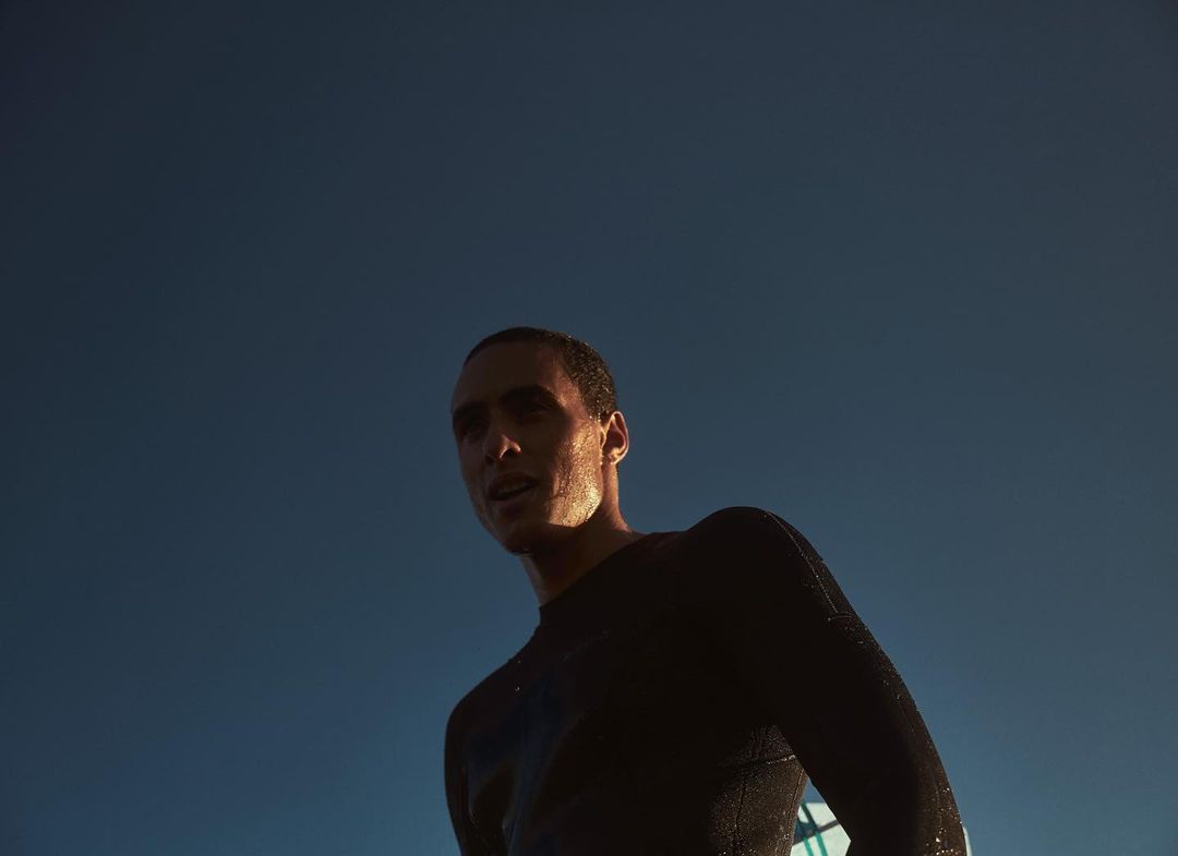 Low-angle shot of a male athlete in dark activewear against a blue sky. Golden light highlights his face and the texture of his clothing.