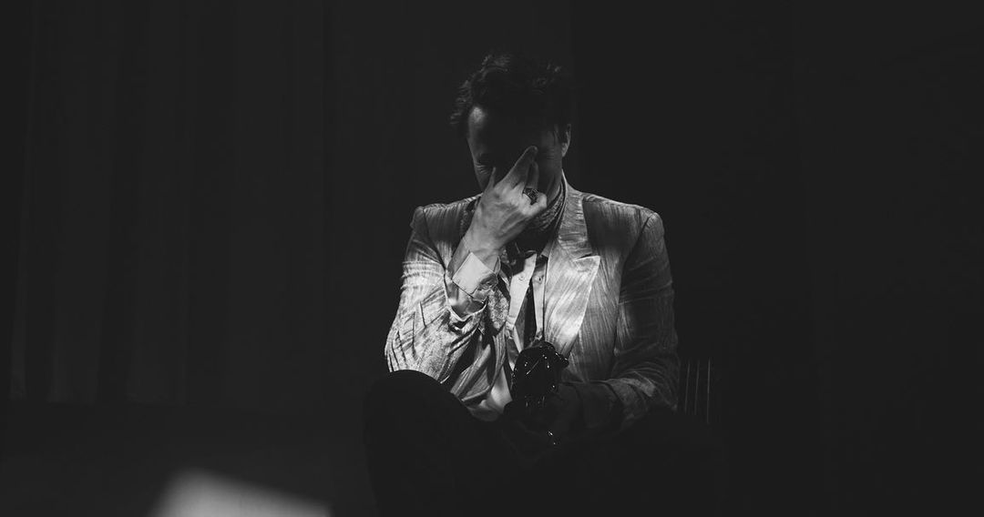 Black and white portrait of a man in a patterned blazer sitting in a dark setting, covering his face with one hand, holding an object.