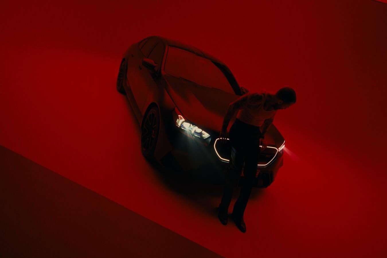 A shirtless man stands in front of a dark luxury sedan under dramatic red lighting, creating a striking automotive advertisement.