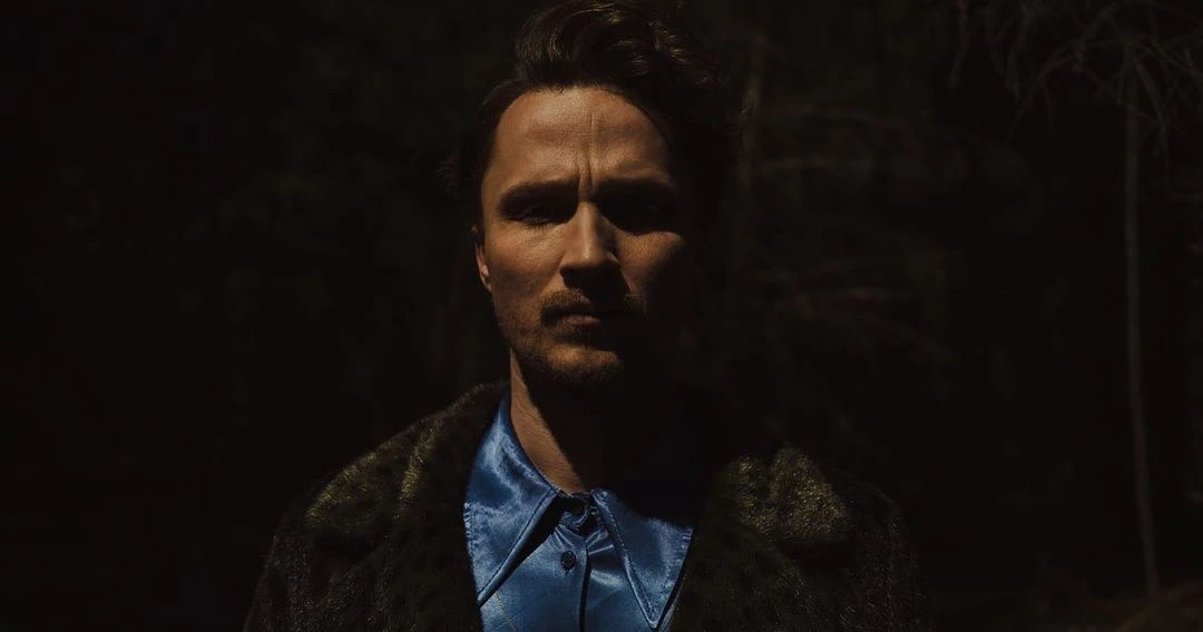 A man in a blue shirt and green coat stares intensely at the camera, illuminated against a dark outdoor backdrop.
