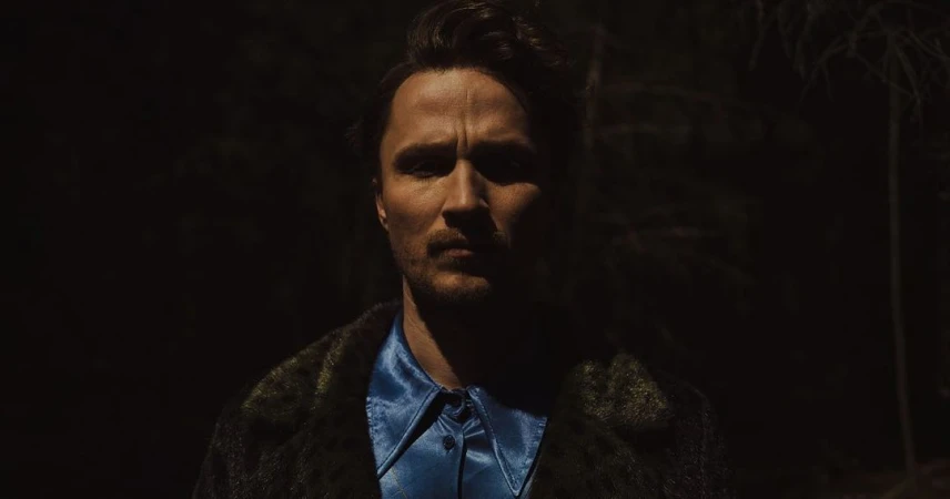 A man in a blue shirt and green coat stares intensely at the camera, illuminated against a dark outdoor backdrop.