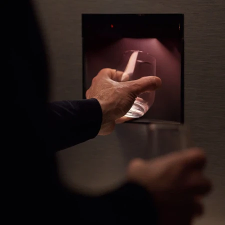 Close-up shot of a person's hand holding a glass filled with water in front of a dispenser, emphasizing hydration and accessibility.
