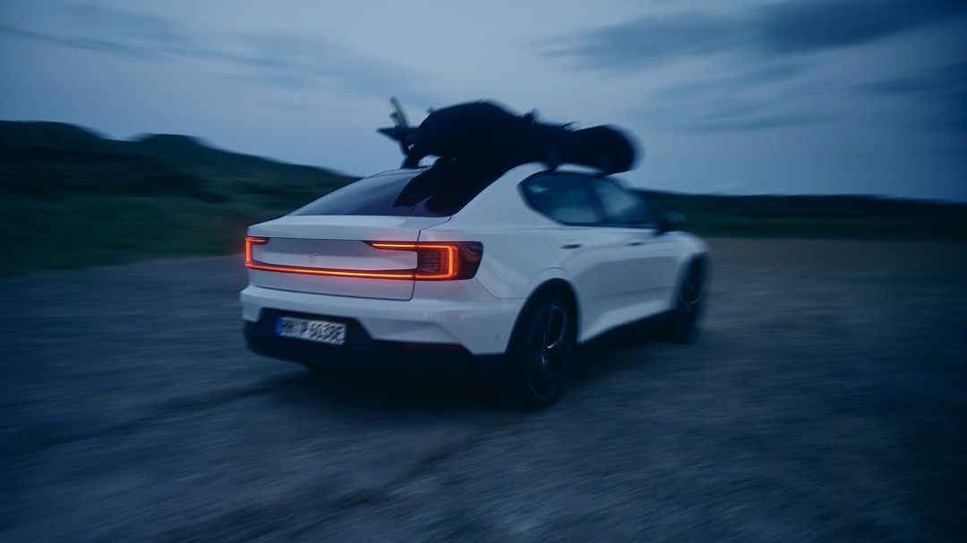 White electric car drives on a road with luggage on the roof rack, against a backdrop of twilight sky and rolling hills.