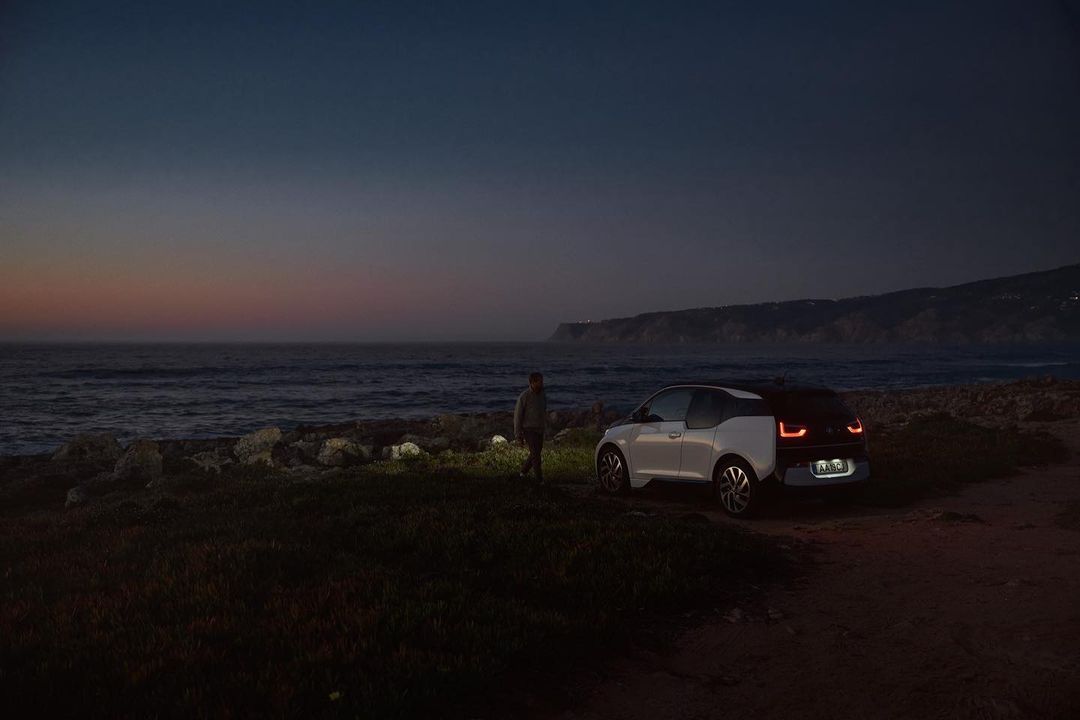 White electric car parks near the ocean with a man standing nearby against a twilight sky at dusk, creating a scenic automotive landscape.