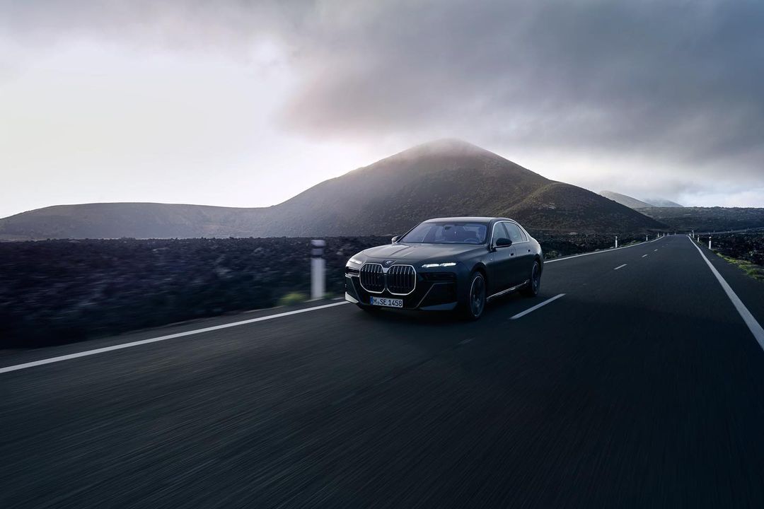 A dark grey BMW 7 Series driving on a dark asphalt road with a dormant volcano and cloudy sky in the background.