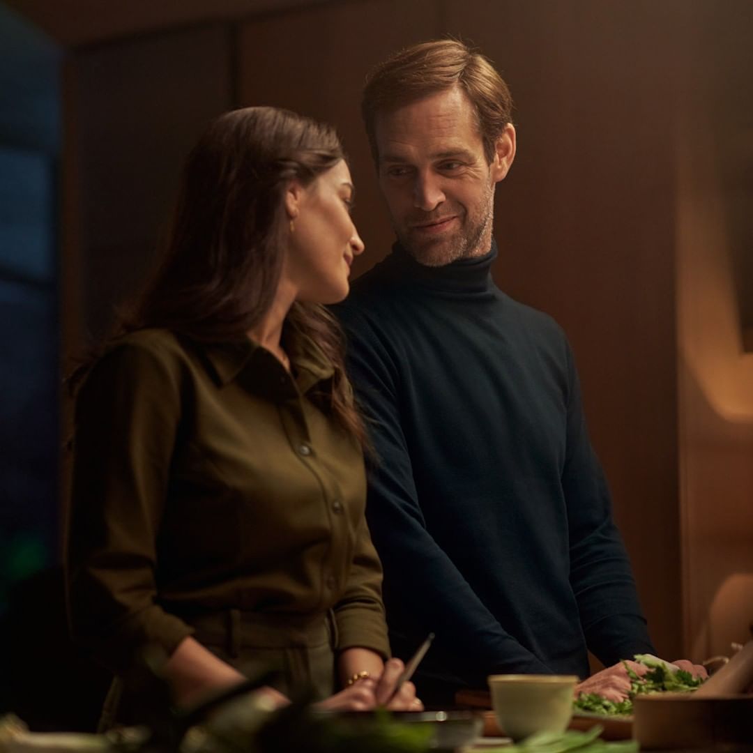 Indoor medium shot of an adult couple with fair skin standing together, wearing fashionable clothing while preparing food at a table.