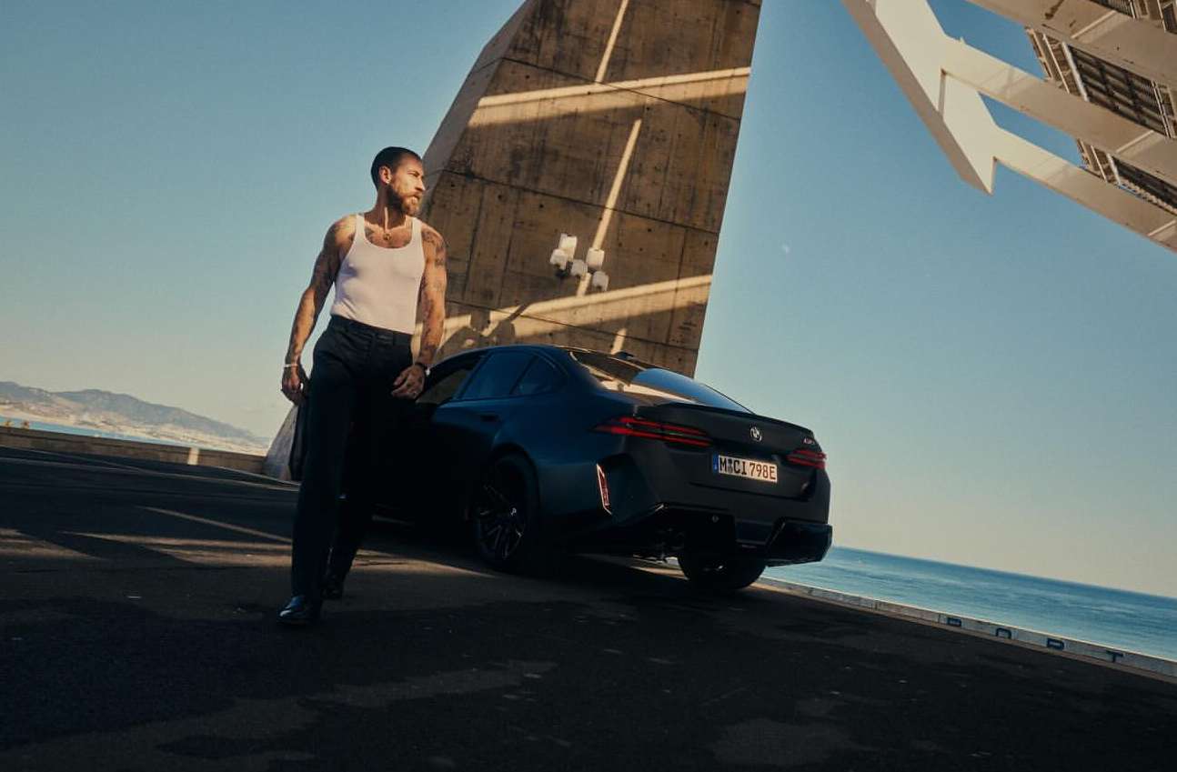 Dark blue BMW sedan parked on asphalt with an adult man in white tank top, concrete architecture, and sea background on clear day.
