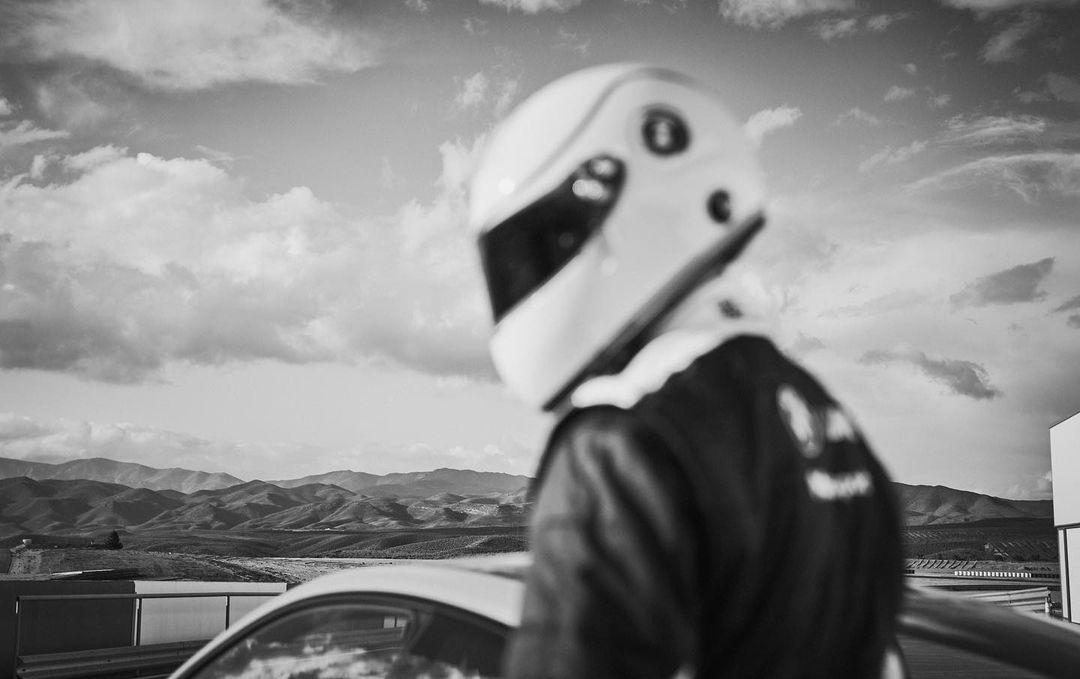 Person in a racing helmet and jacket leans against a car at a racetrack with mountains and cloudy sky visible in black and white.
