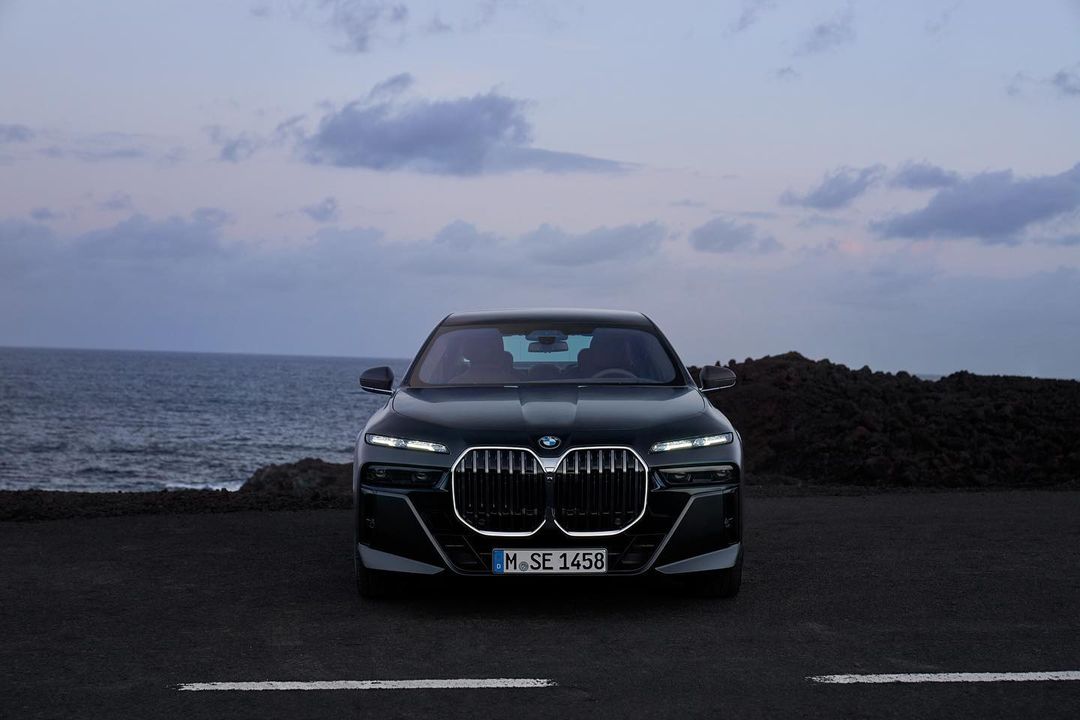 Front view of a dark green BMW 7 Series parked on a black asphalt surface, against a seascape and dusk sky background.