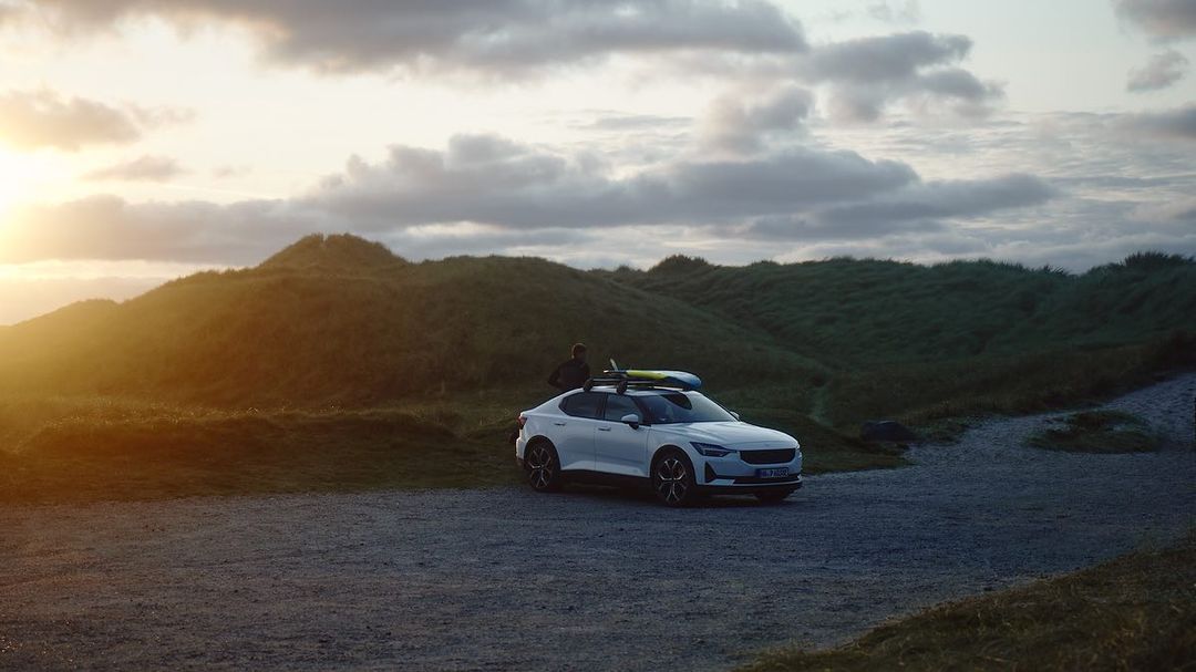 White sportscar parked on a gravel road near grassy dunes under a cloudy sky with a surfboard strapped to the roof.