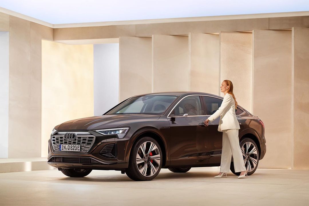 Brown Audi Q8 e-tron electric car in a studio setting, woman in a white pantsuit opening the driver's side door, neutral background.