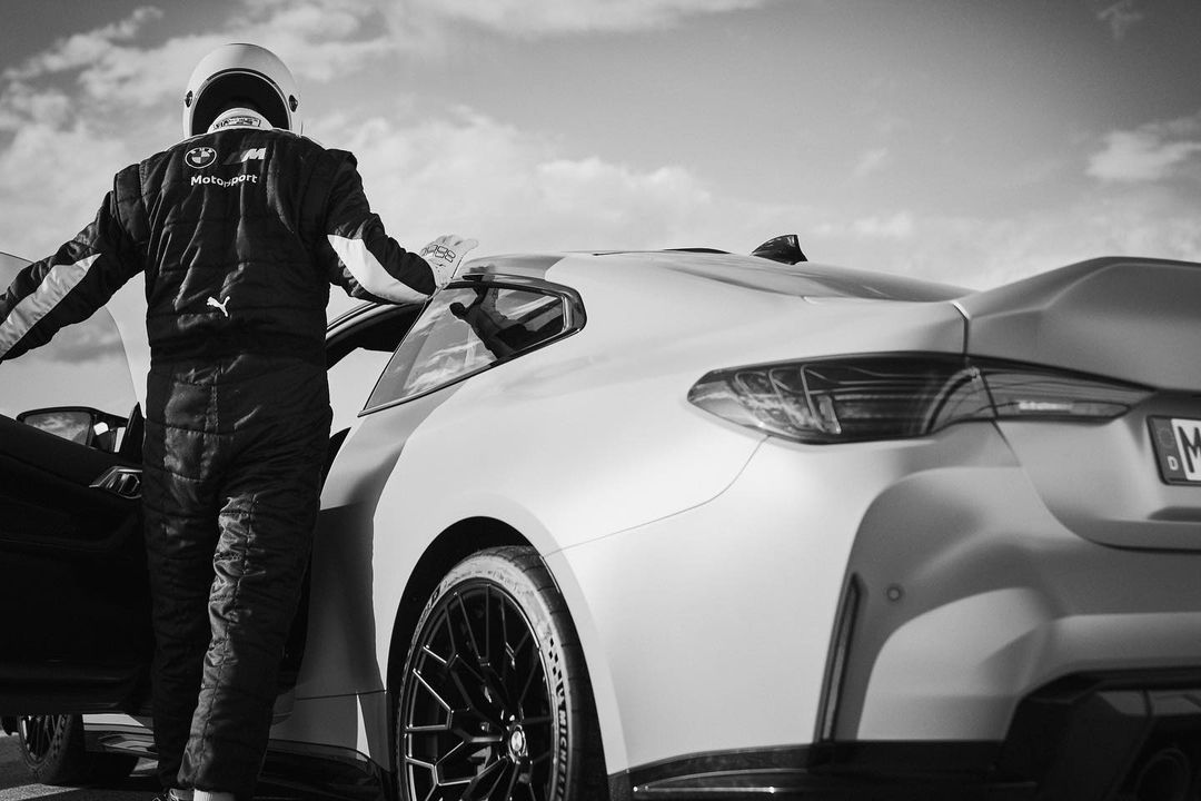 Monochrome shot of race car driver in uniform and helmet standing near BMW race car on track, showcasing automotive design and racing elements.