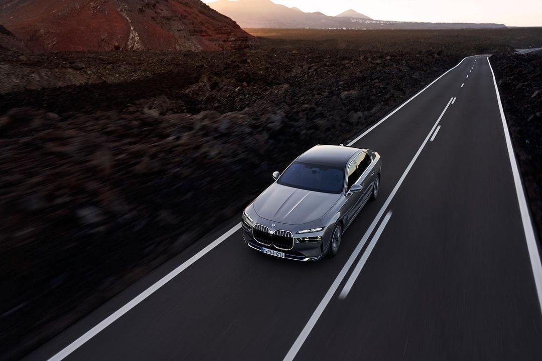 Grey BMW 7 Series sedan driving on an asphalt road through a rocky landscape with a mountain in the background at dusk.