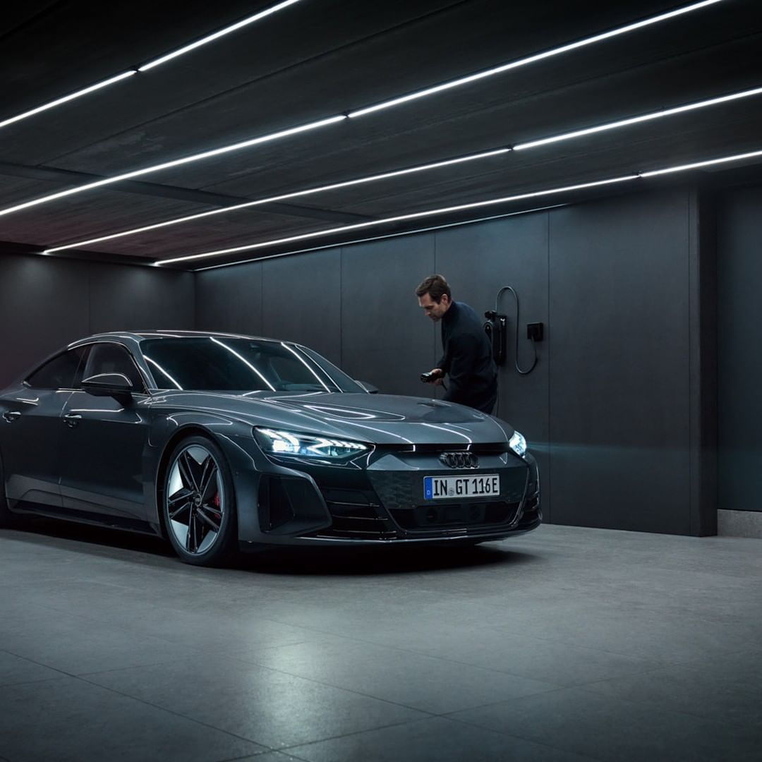 Gray Audi e-tron GT parked indoors, with an adult man near a wall-mounted charger, under bright white ceiling lights.