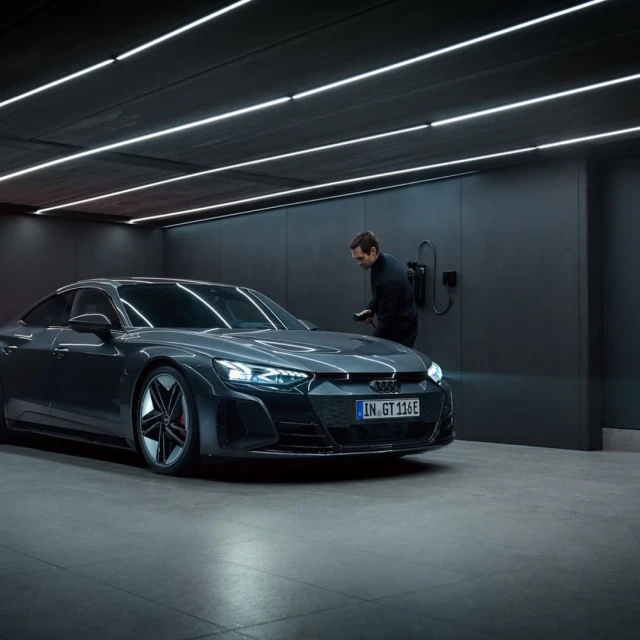 Gray Audi e-tron GT parked indoors, with an adult man near a wall-mounted charger, under bright white ceiling lights.