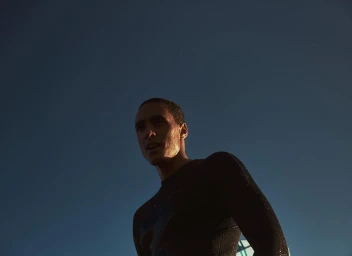 Man wearing a dark wetsuit with water droplets on his skin against a clear blue sky background