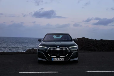 Front view of a dark luxury sedan parked on a coastal road at dusk under a cloudy sky, captured by Manuel Nagel.
