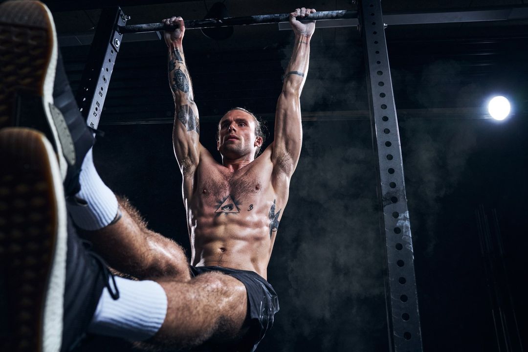Muscular man with tattoos does pull-ups on a bar in a dark, smoky gym, showing his abs and arm muscles under a bright overhead light.