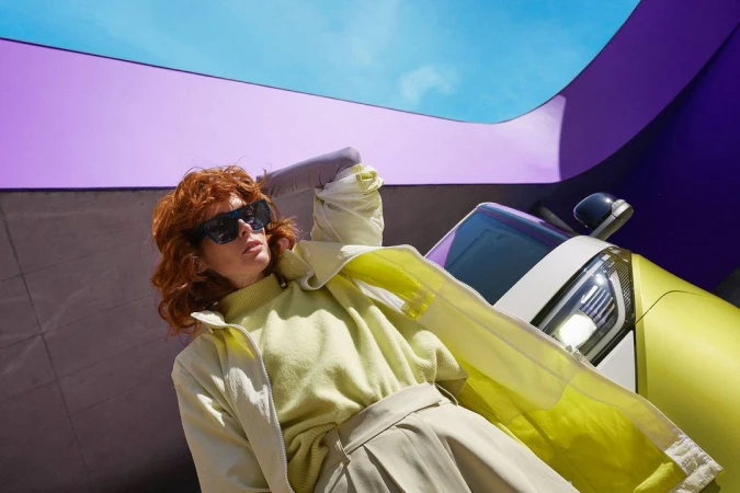 Red-haired woman wearing sunglasses and a pale yellow outfit posing outdoors next to a modern car under purple architectural structures