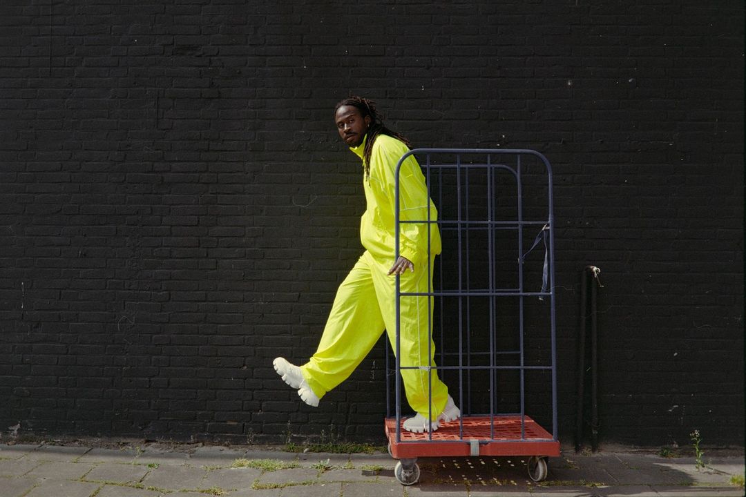 An adult Black male wearing a neon tracksuit and white sneakers stands next to a metal utility cart against a dark brick wall outdoors.