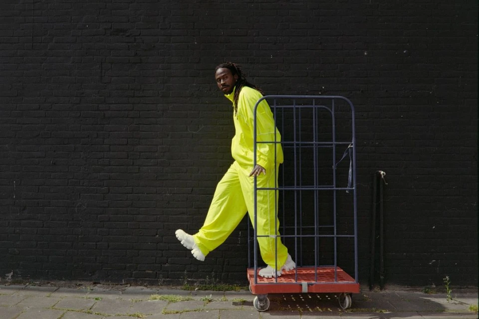 An adult Black male wearing a neon tracksuit and white sneakers stands next to a metal utility cart against a dark brick wall outdoors.
