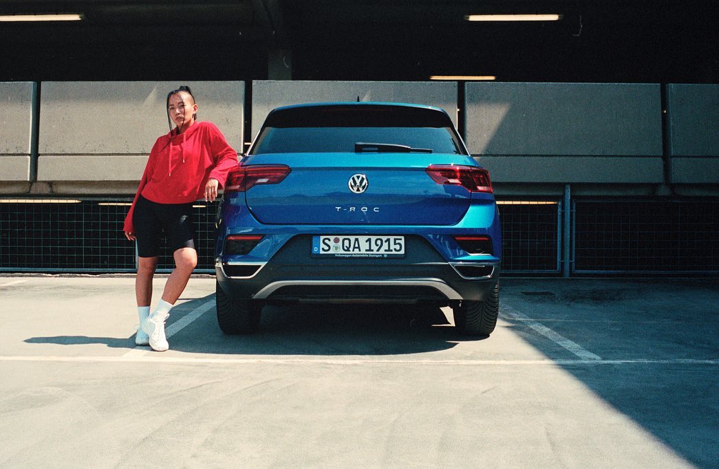 A woman in a red hoodie poses beside a blue Volkswagen T-Roc in an urban parking garage, capturing a blend of fashion and automotive style.