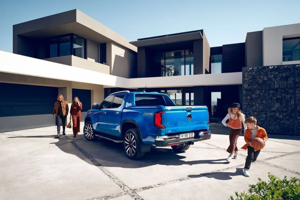Family next to a blue Volkswagen Amarok pickup truck in the driveway of a modern house on a sunny day; automotive lifestyle.