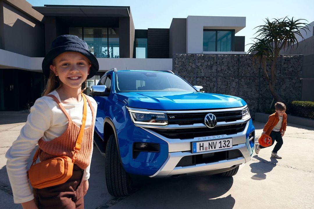 A blue Volkswagen Amarok pickup truck parked outside a modern house with a girl in the foreground and a boy playing nearby.