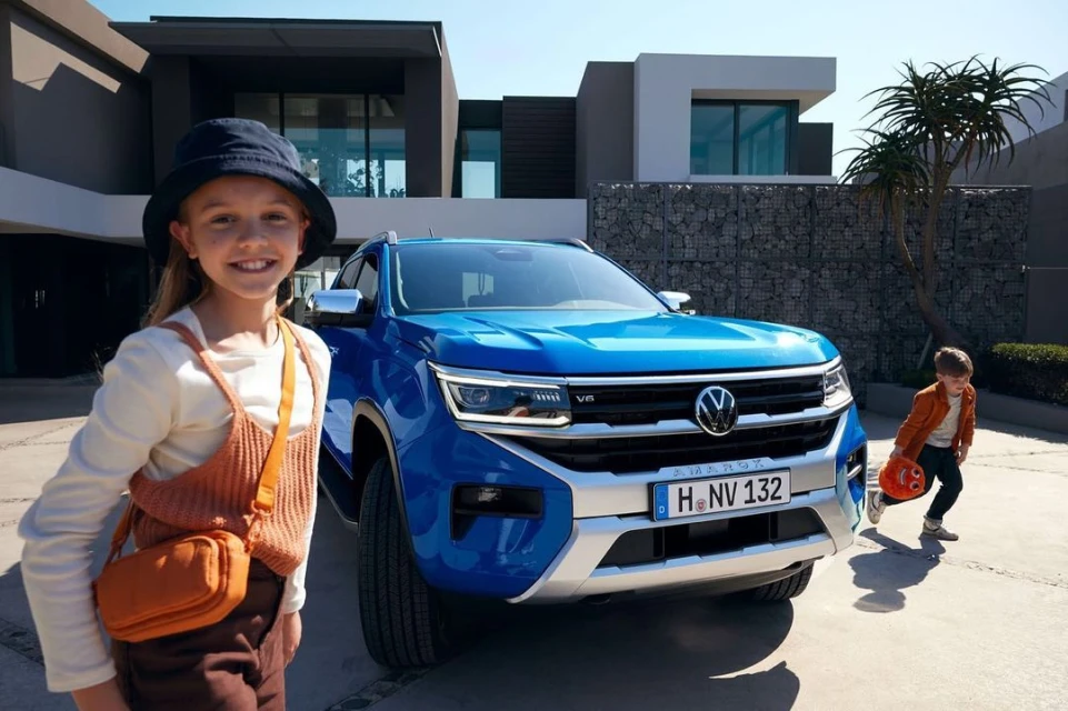 A blue Volkswagen Amarok pickup truck parked outside a modern house with a girl in the foreground and a boy playing nearby.