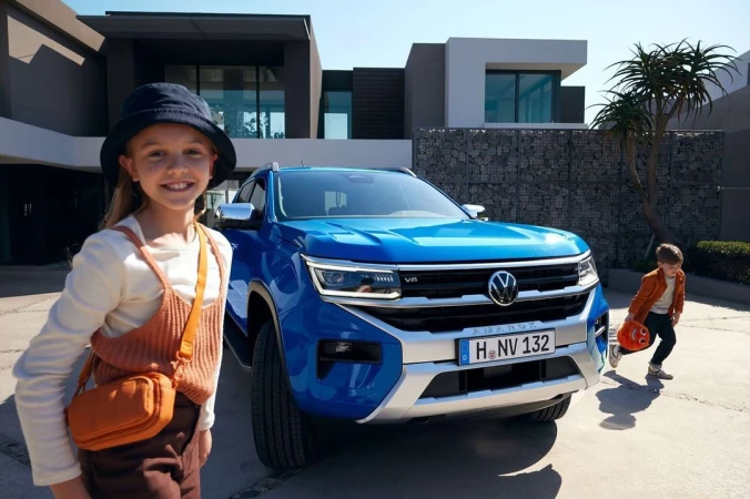 A blue Volkswagen Amarok pickup truck parked outside a modern house with a girl in the foreground and a boy playing nearby.