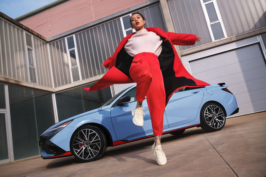 Woman in red suit poses energetically with blue Hyundai car against modern building for automotive and fashion lifestyle visual content.