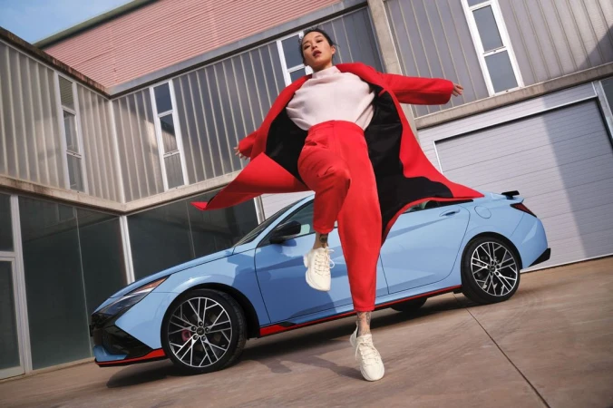 Woman in red suit poses energetically with blue Hyundai car against modern building for automotive and fashion lifestyle visual content.