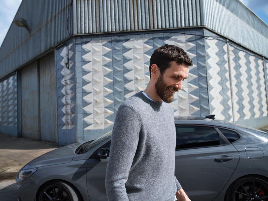 A man smiles next to a gray car in front of a textured building, capturing a casual, candid moment in natural light.