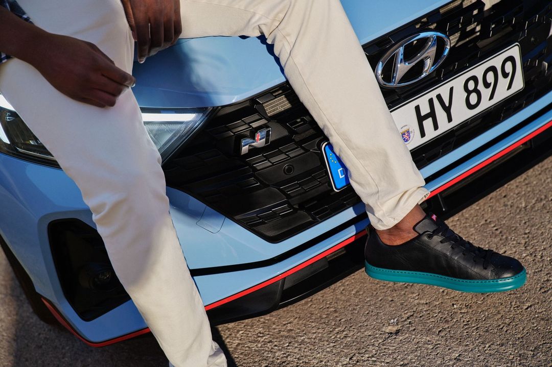 A man in cream pants sits on the hood of a light blue Hyundai car, his black sneaker resting on the vehicle's front grill.