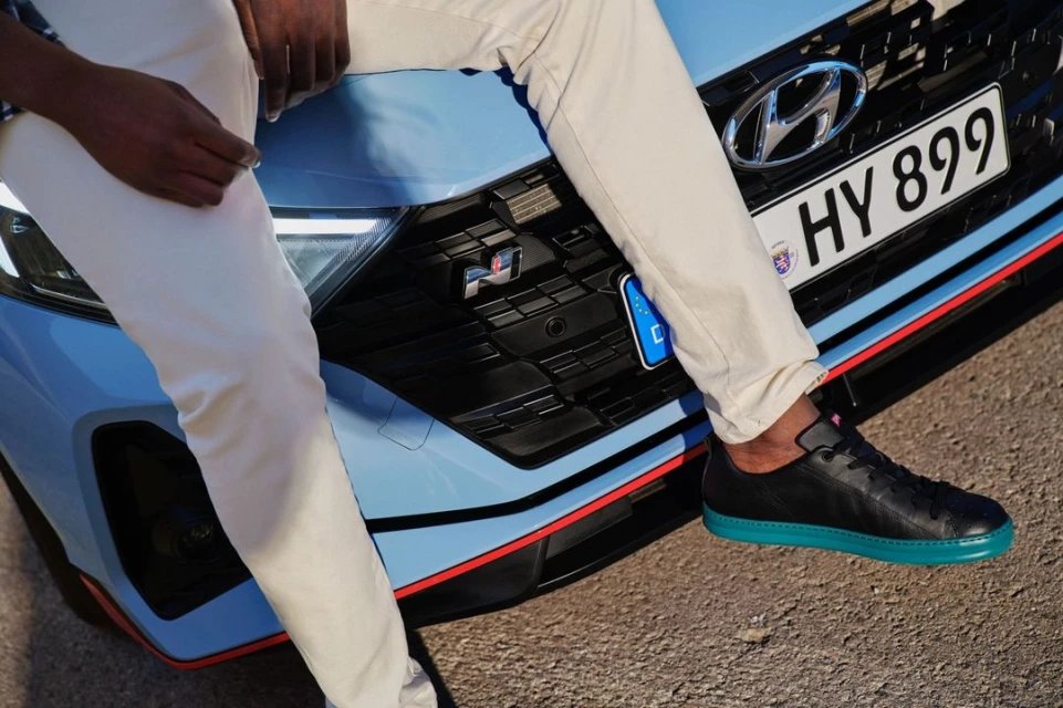 A man in cream pants sits on the hood of a light blue Hyundai car, his black sneaker resting on the vehicle's front grill.