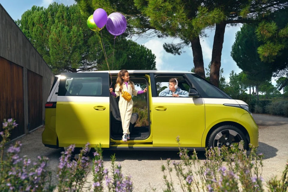 A yellow and white Volkswagen ID. Buzz with two kids and balloons, parked outdoors near trees and foliage, showcasing a family travel scene.